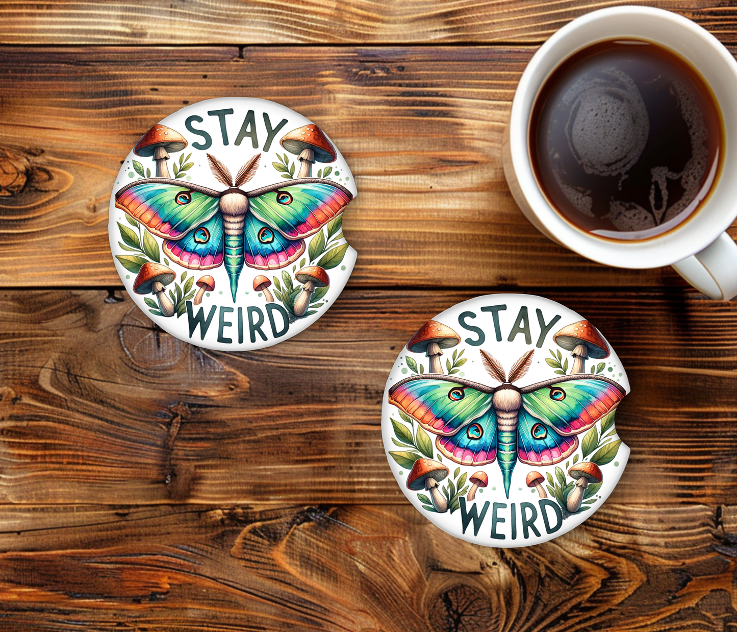 Two colorful butterfly coasters with 'Stay Weird' text on a wooden surface with a cup of coffee.