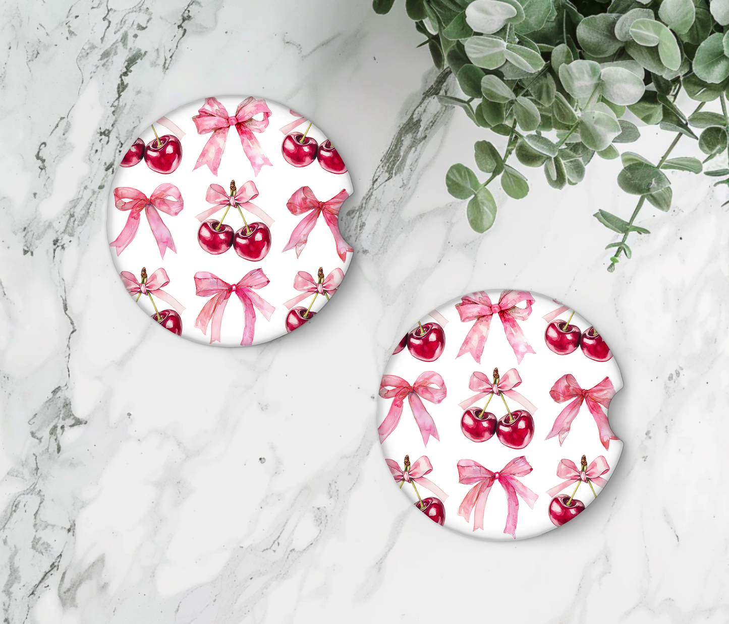 Two round coasters with cherry and bow design on a marble surface with greenery.