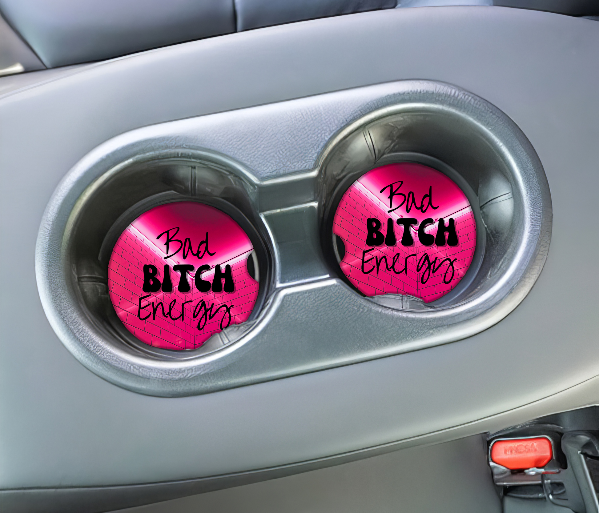 Two pink car coasters with 'Bad Bitch Energy' text in a car's cup holder.