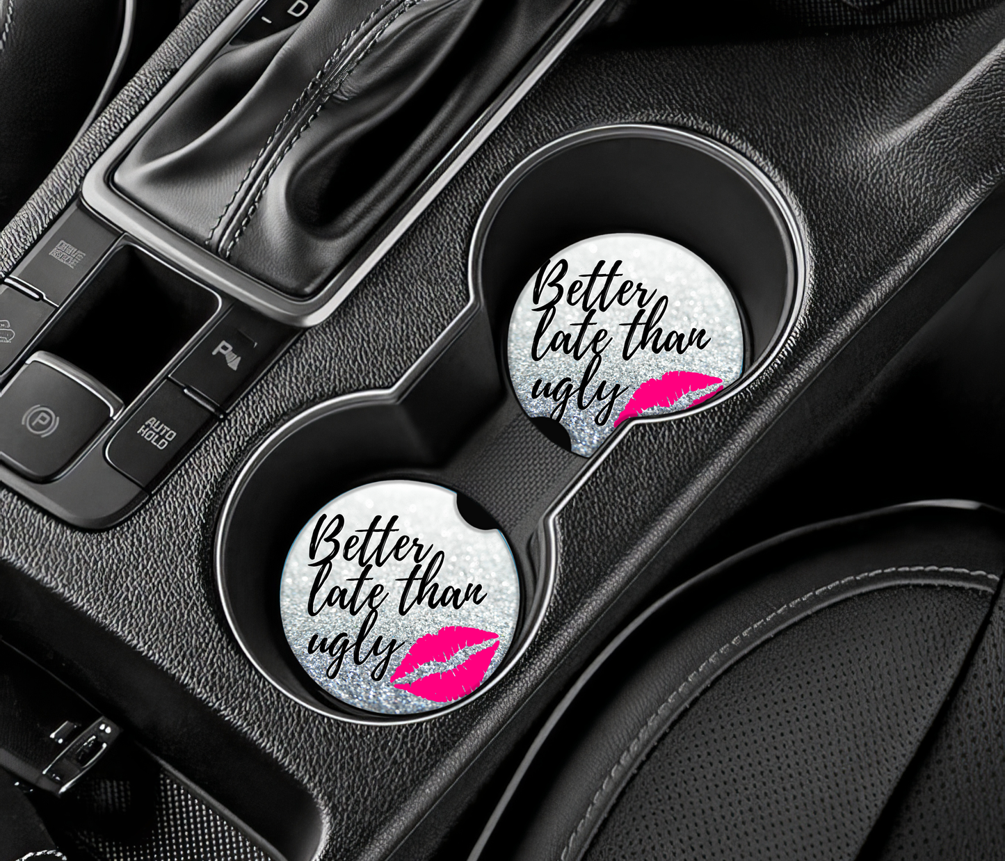 Two car coasters with 'Better late than ugly' text and pink lip print on a car cup holder.