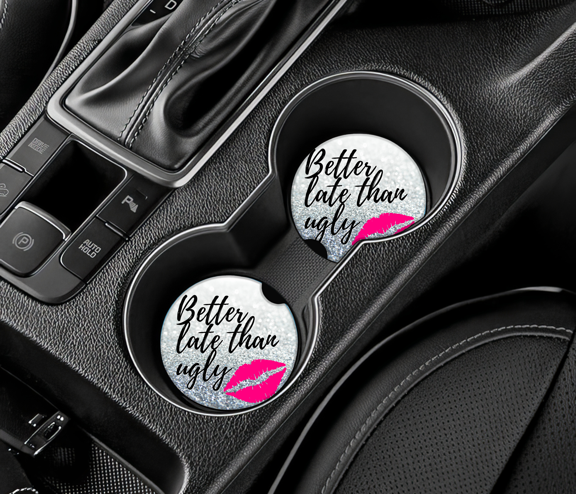 Two car coasters with 'Better late than ugly' text and pink lip print on a car cup holder.