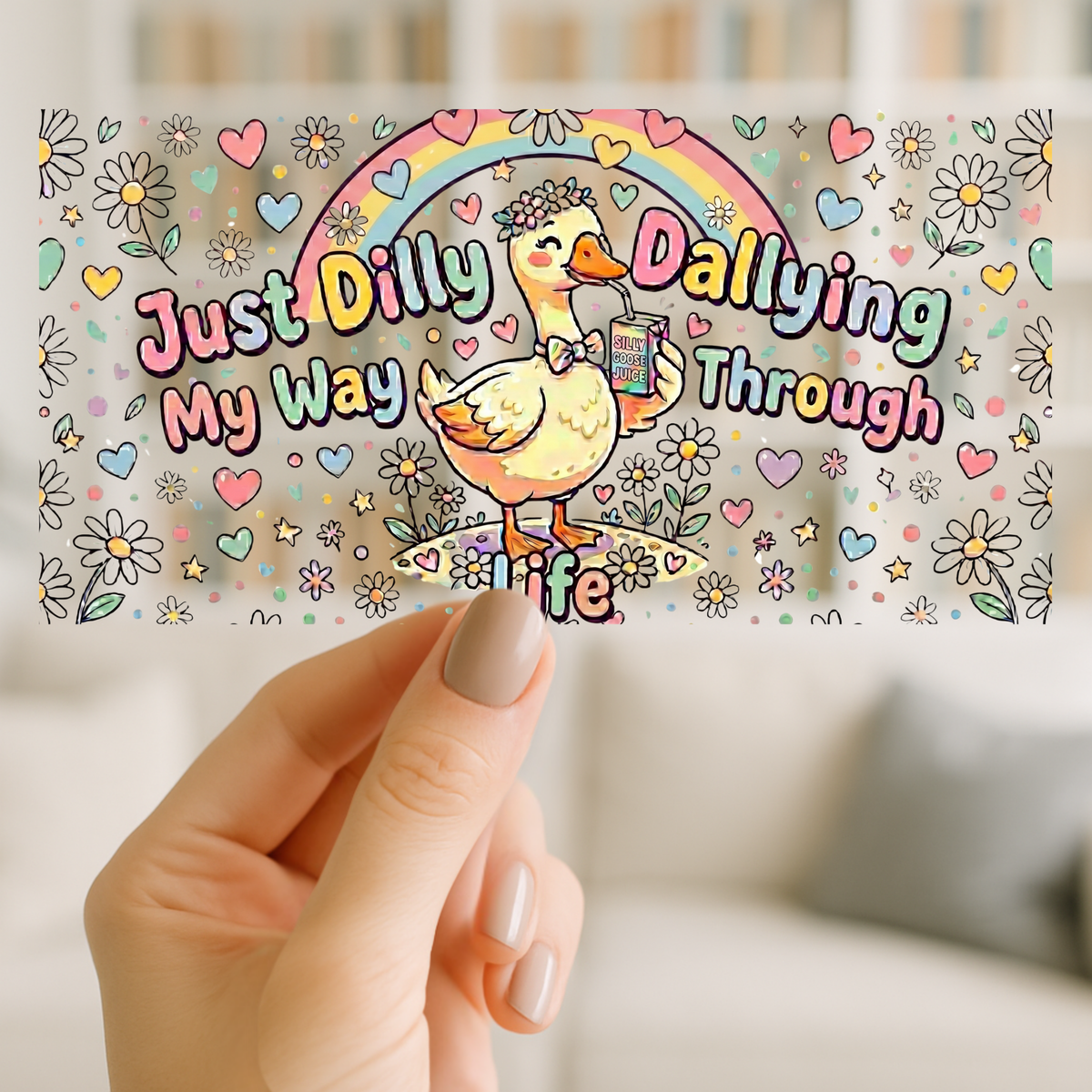 A hand holds a rectangular sticker or card featuring a colorful cartoon duck under a rainbow with the phrase 'Just Dilly Dallying My Way Through Life' on a softly blurred indoor background.