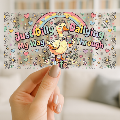 A hand holds a rectangular sticker or card featuring a colorful cartoon duck under a rainbow with the phrase 'Just Dilly Dallying My Way Through Life' on a softly blurred indoor background.